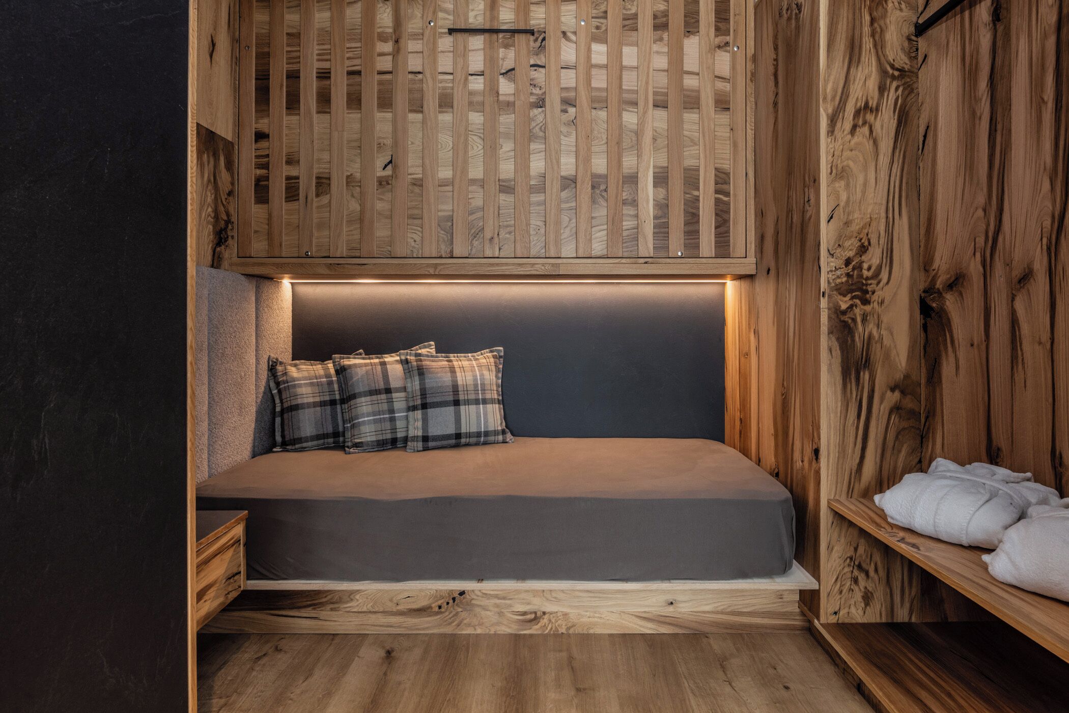 A cozy room with a wooden slat ceiling, a bed with grey bedding and plaid pillows, and a shelf with towels.