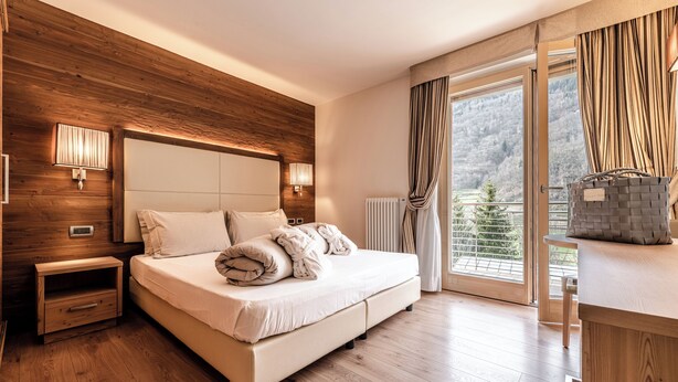 Ein modernes Schlafzimmer mit einem hölzernen Kopfteil, einem großen Bett, Nachttischlampen, einem Nachttisch aus Holz und Blick auf einen Wald durch ein großes Fenster.