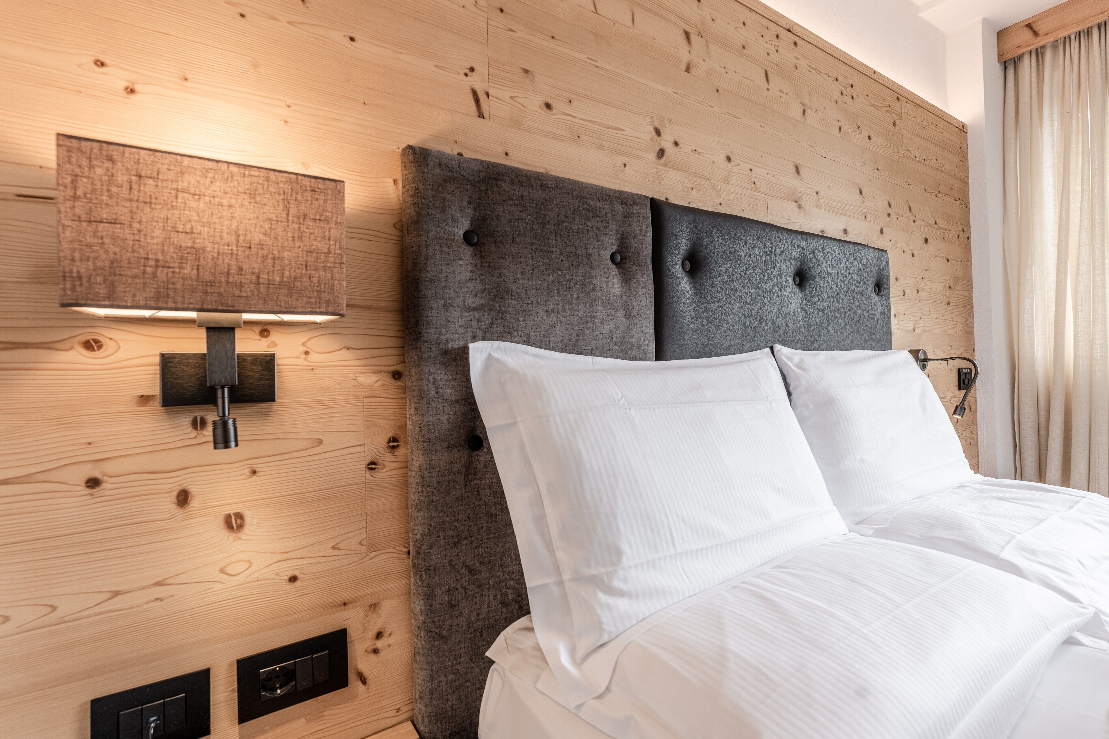 A neatly made bed with white linens, a dark upholstered headboard, and a wall-mounted light fixture.