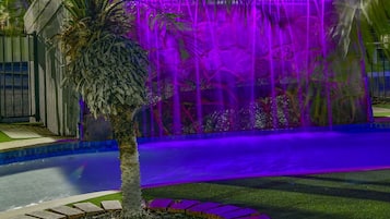 Un espacio al aire libre iluminado con luces moradas, con una fuente de agua, rodeado de palmeras y un camino pavimentado.