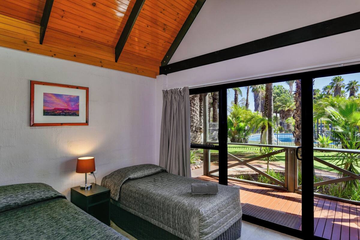 Quarto com cama, mesa de cabeceira com abajur, quadro na parede, e vista para uma piscina e palmeiras através de uma porta de vidro.