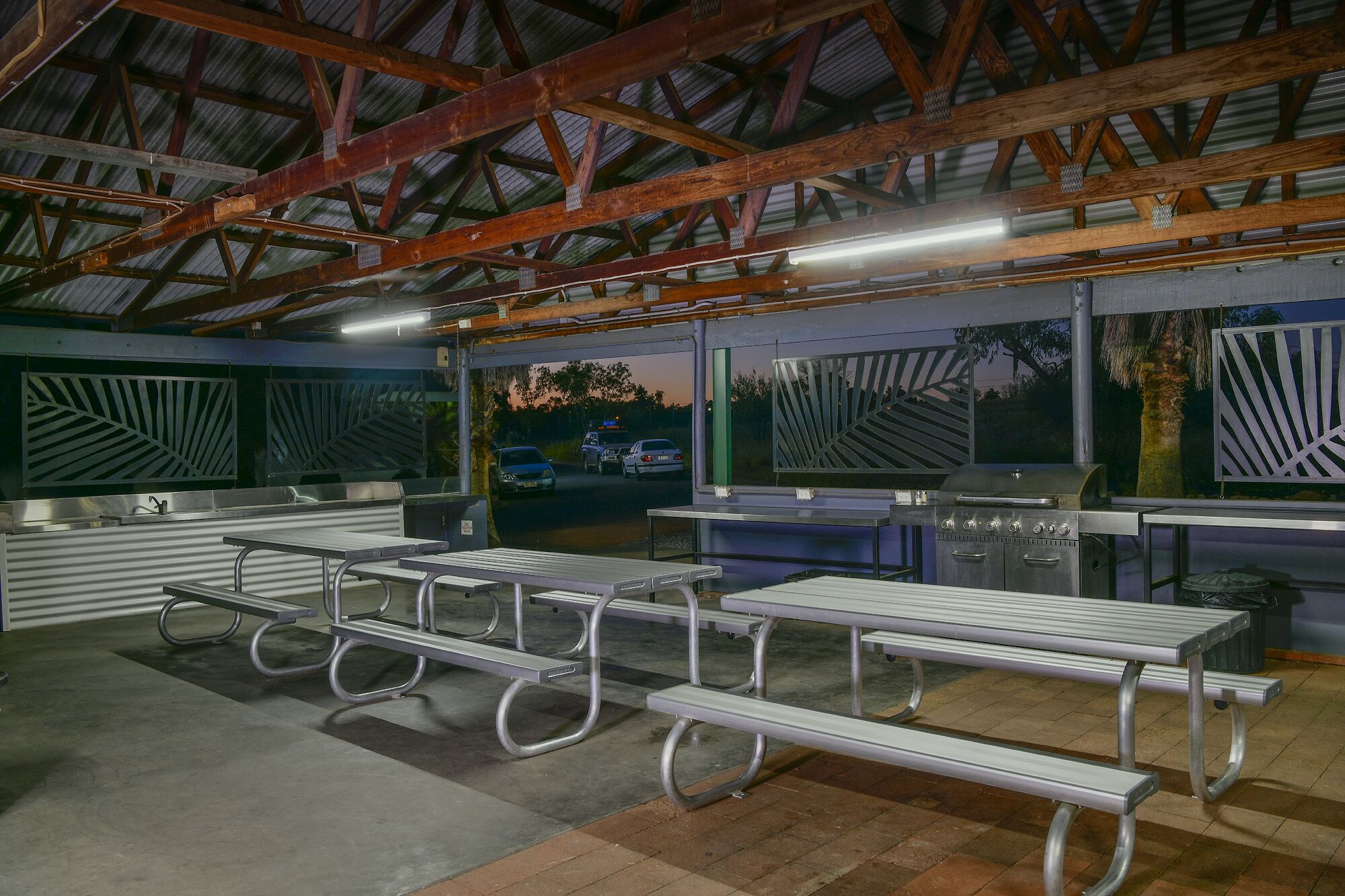 A covered outdoor area with metal picnic tables and a barbecue grill.