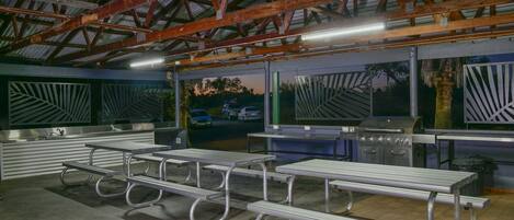A covered outdoor area with metal picnic tables and a barbecue grill.