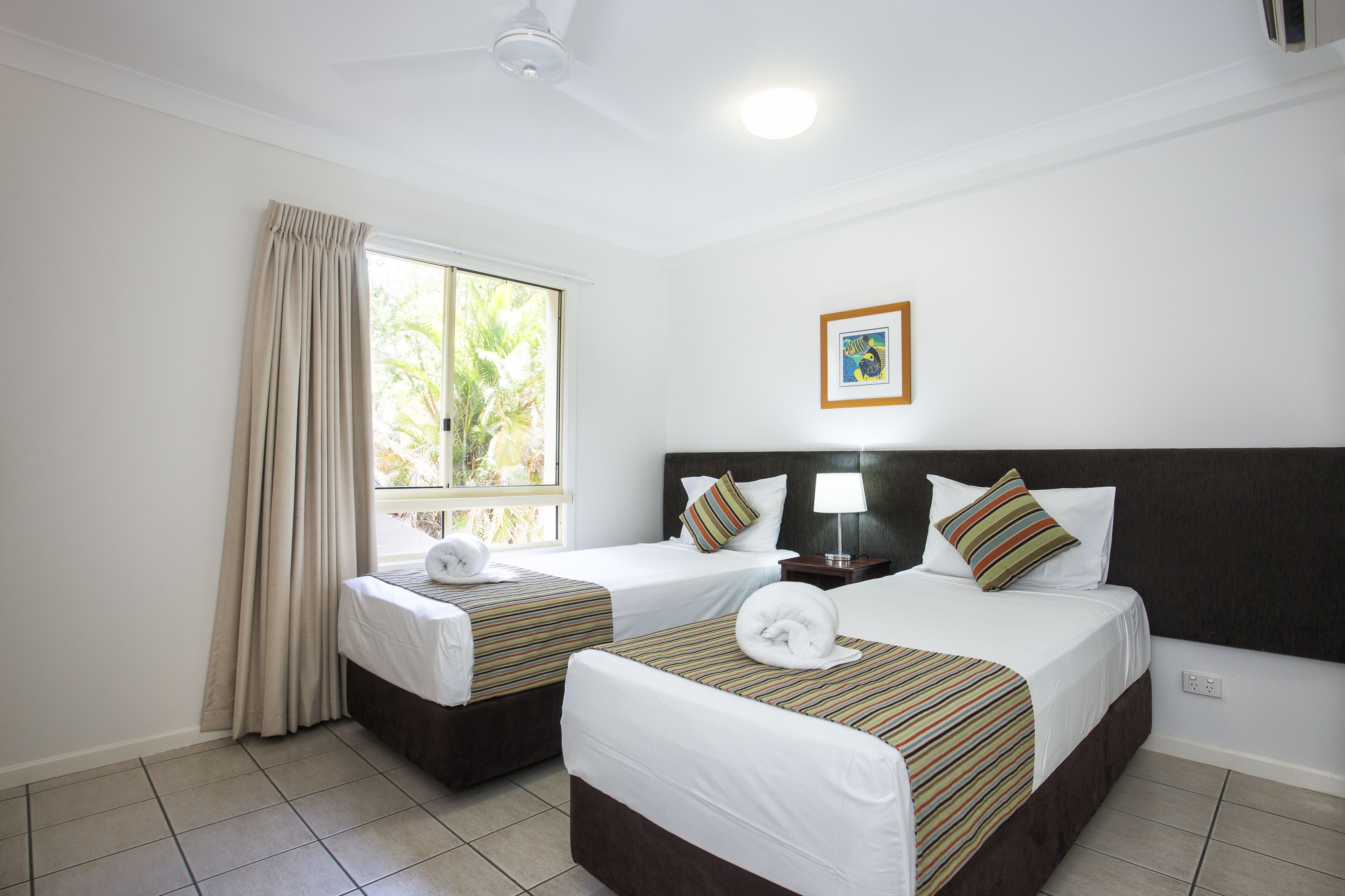 Une chambre d’hôtel avec deux lits, une fenêtre avec des rideaux, un ventilateur de plafond, un tableau encadré au mur et un mur blanc.