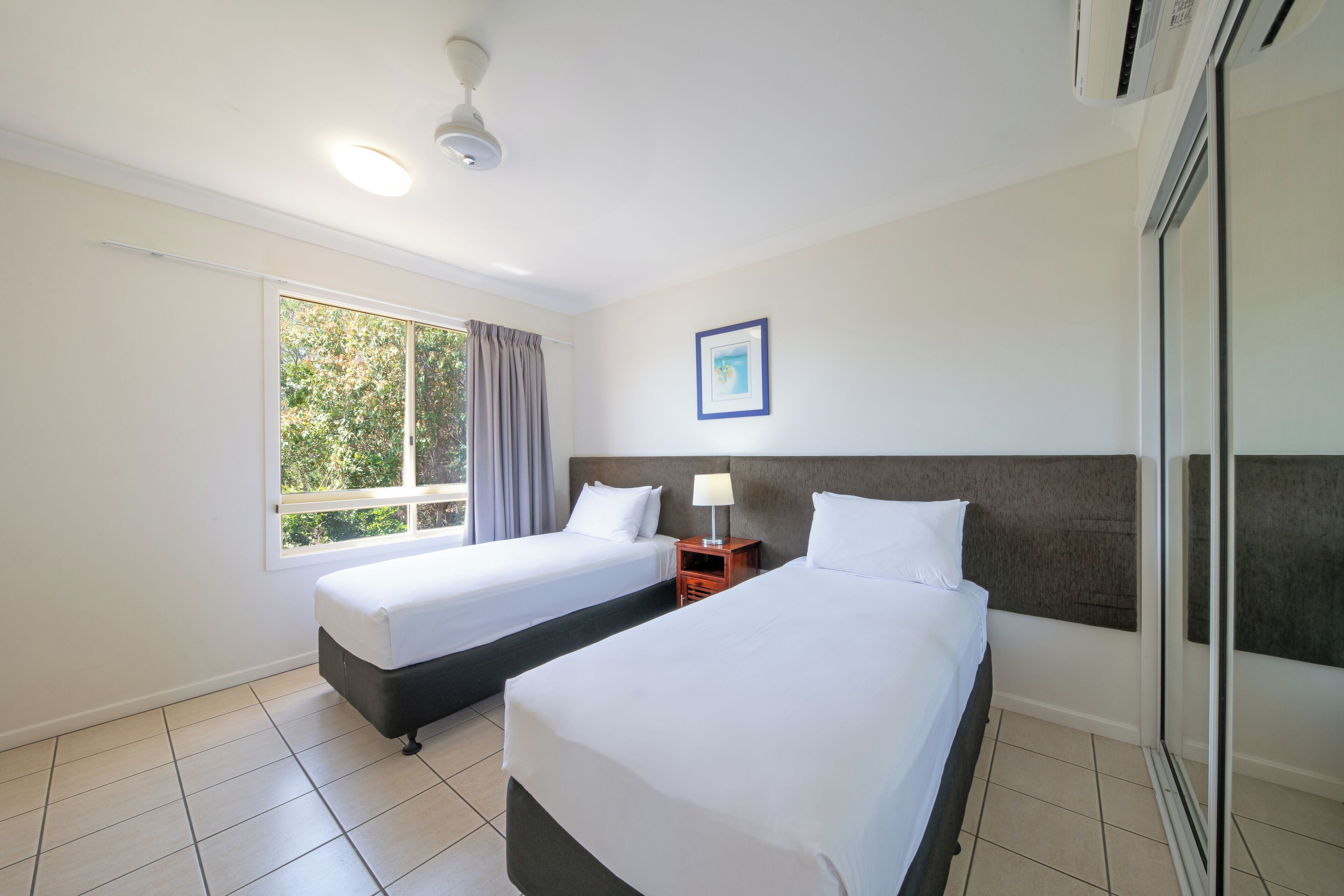 Habitación de hotel con dos camas, una ventana con cortinas, un ventilador de techo y un cuadro en la pared.