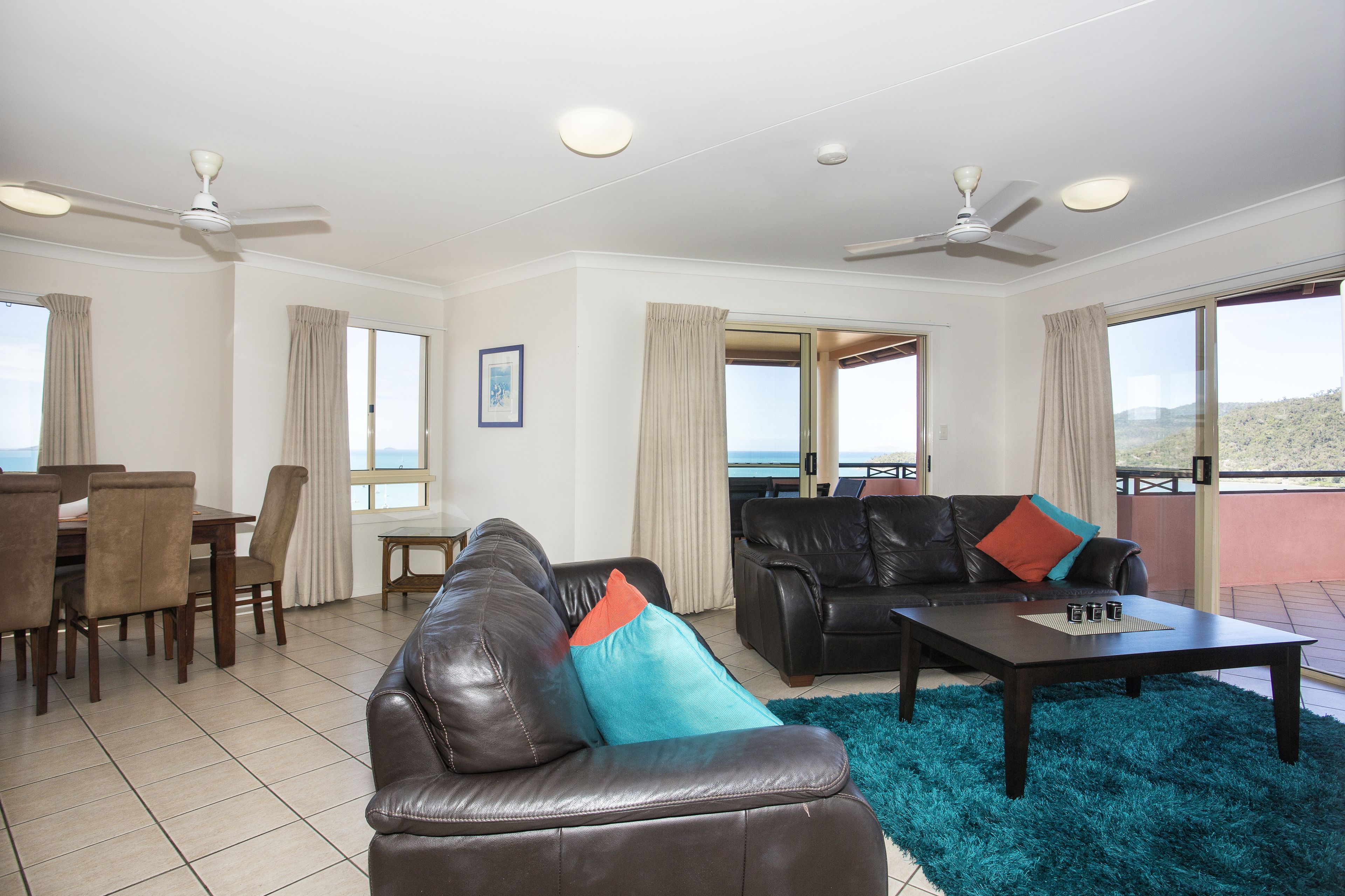 A living room with a leather sofa, a coffee table, a dining area with chairs and a table, and a balcony with a view.