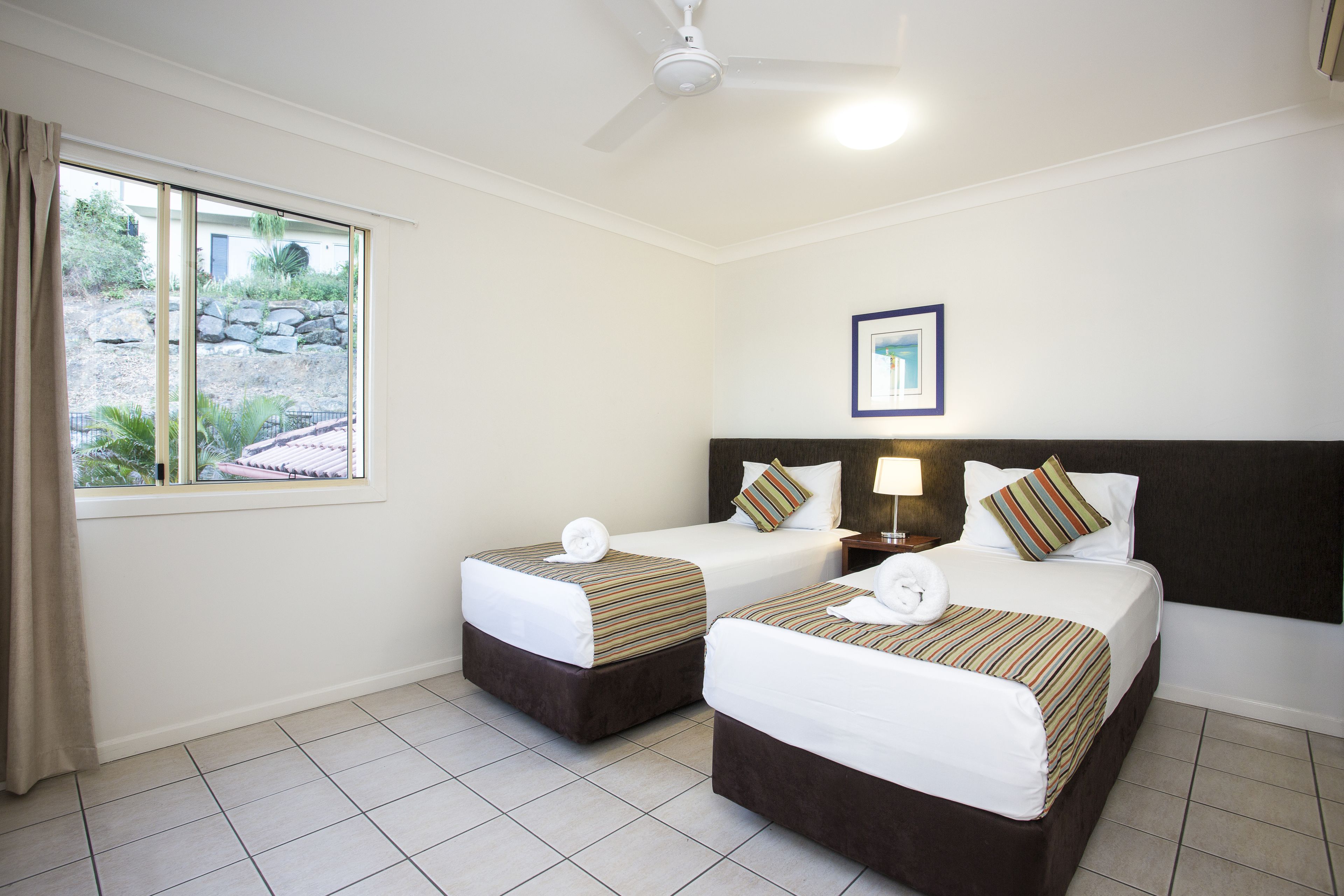 A hotel room with two beds, a ceiling fan, a window with curtains, and a framed picture on the wall.