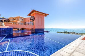 Une piscine à débordement offrant une vue imprenable sur la mer et les bateaux, un bâtiment rose et une terrasse carrelée de bleu.
