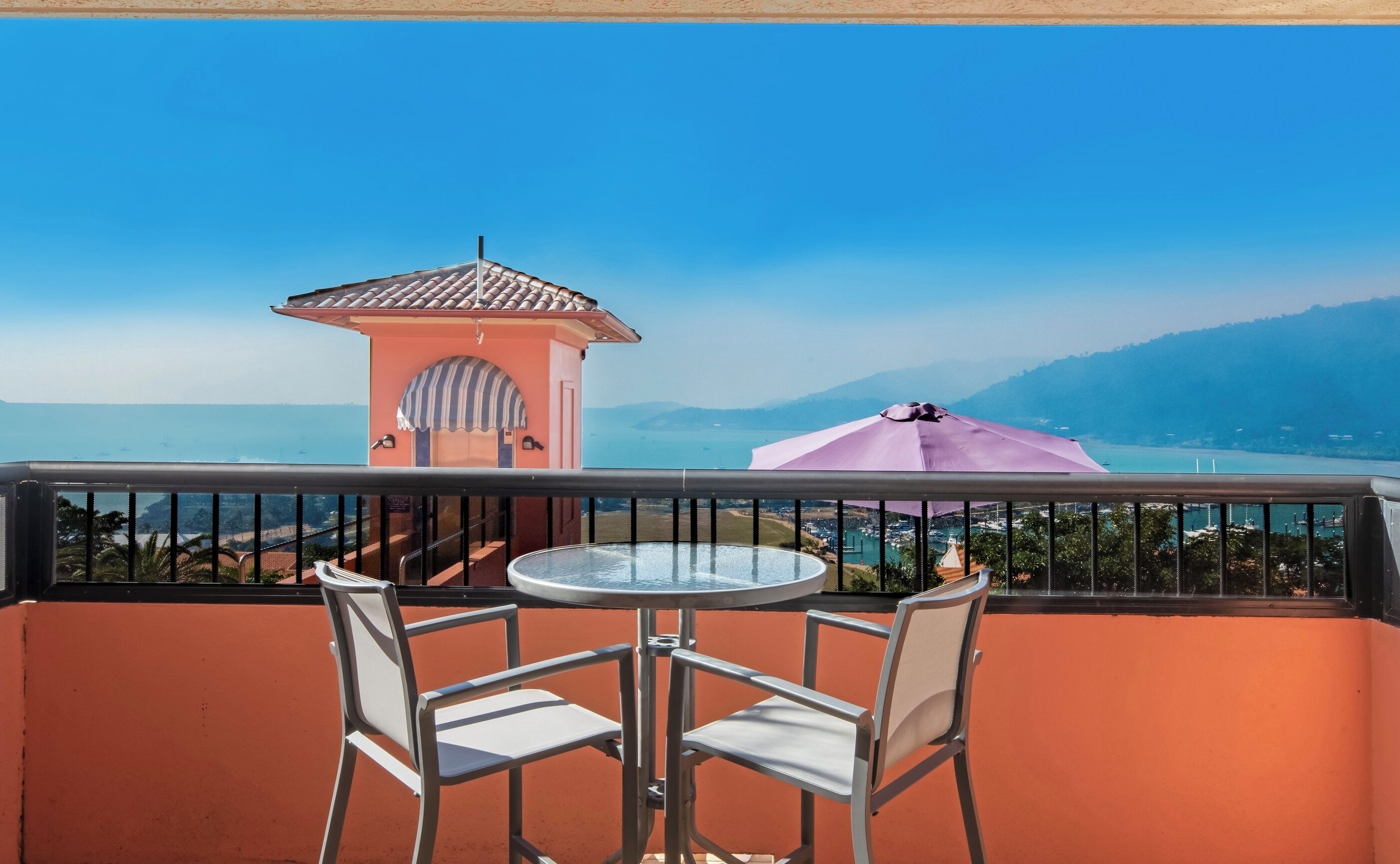 A balcony with a table and chairs, overlooking a coastal view with a small tower and a purple umbrella.