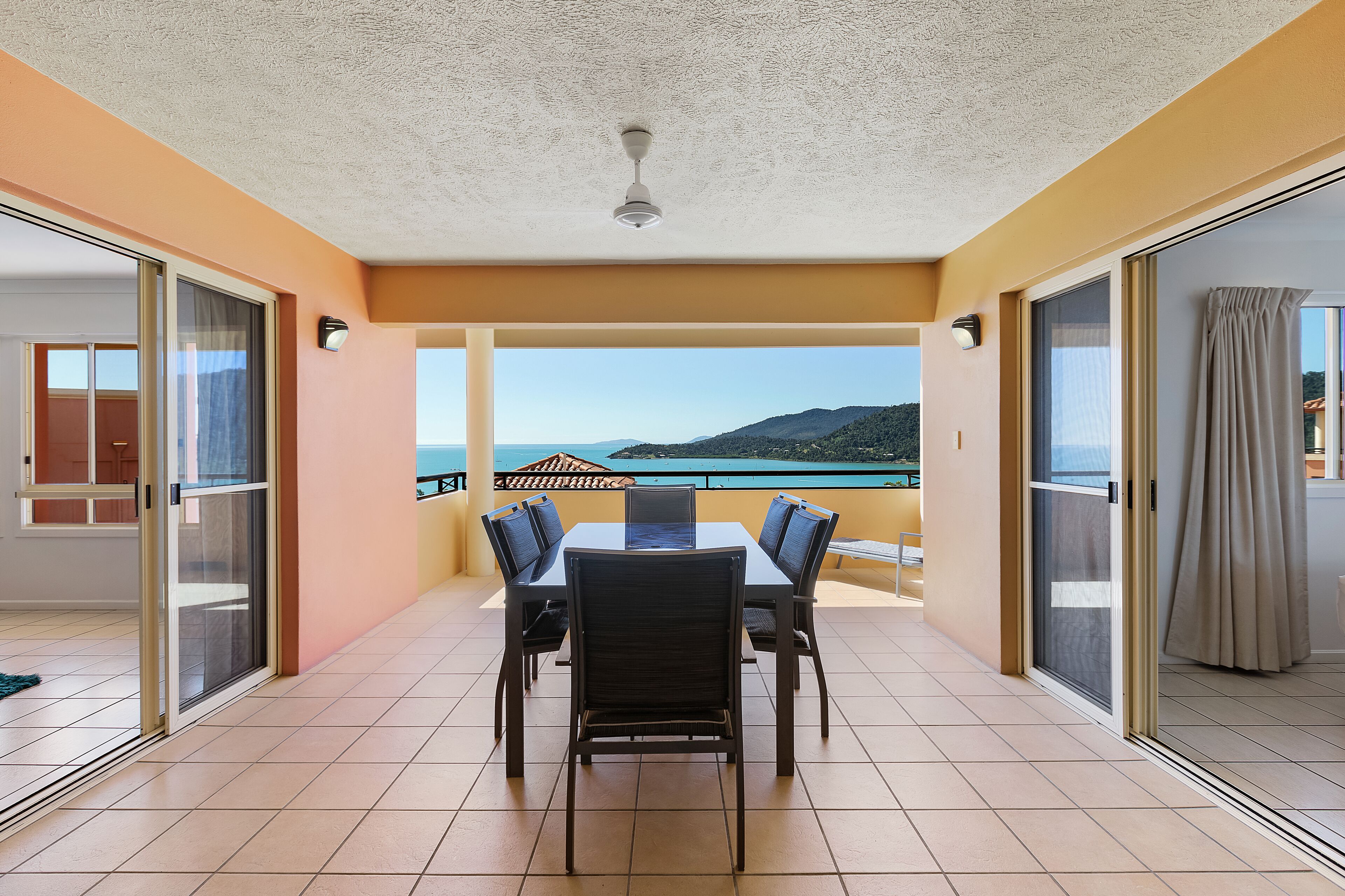 A balcony with a table and chairs, a ceiling fan, and a view of the sea and mountains.