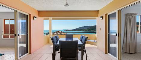 Uma varanda com mesa e cadeiras, um ventilador de teto e vista para o mar e as montanhas.