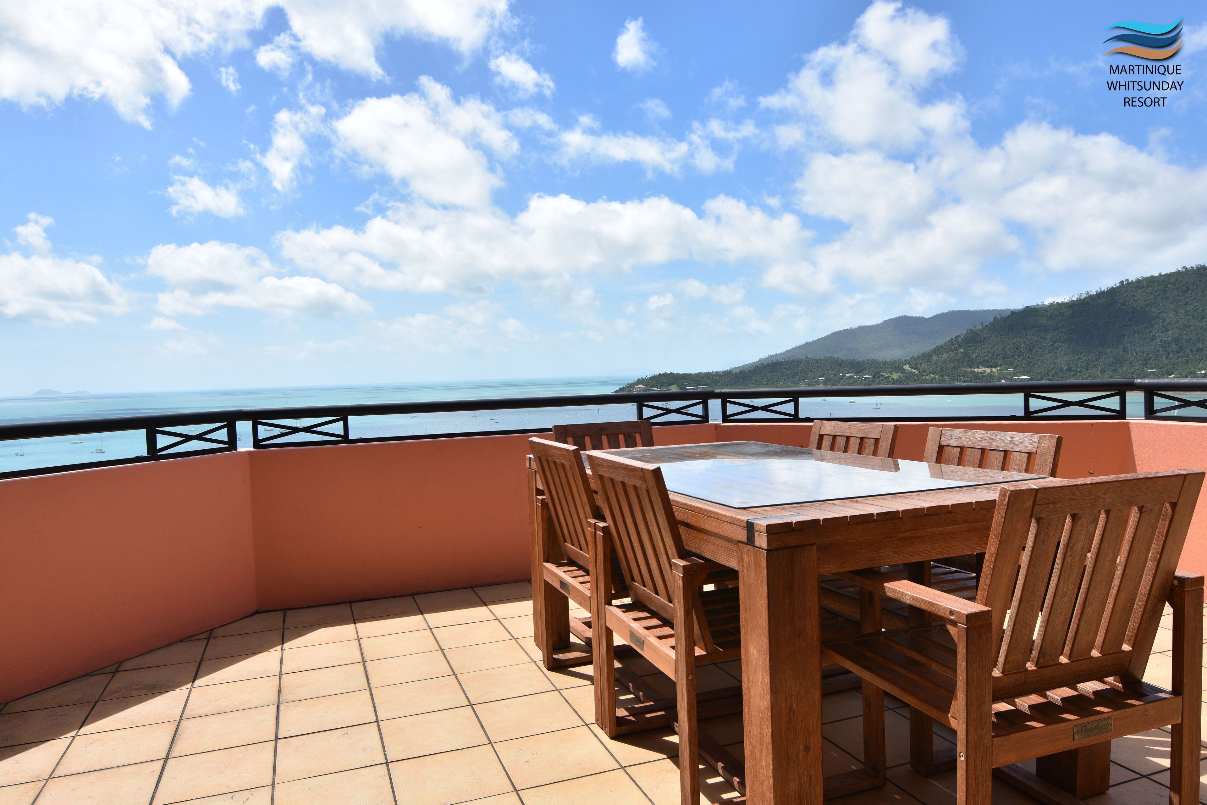 A wooden table and chairs on a balcony with a view of the sea and mountains.