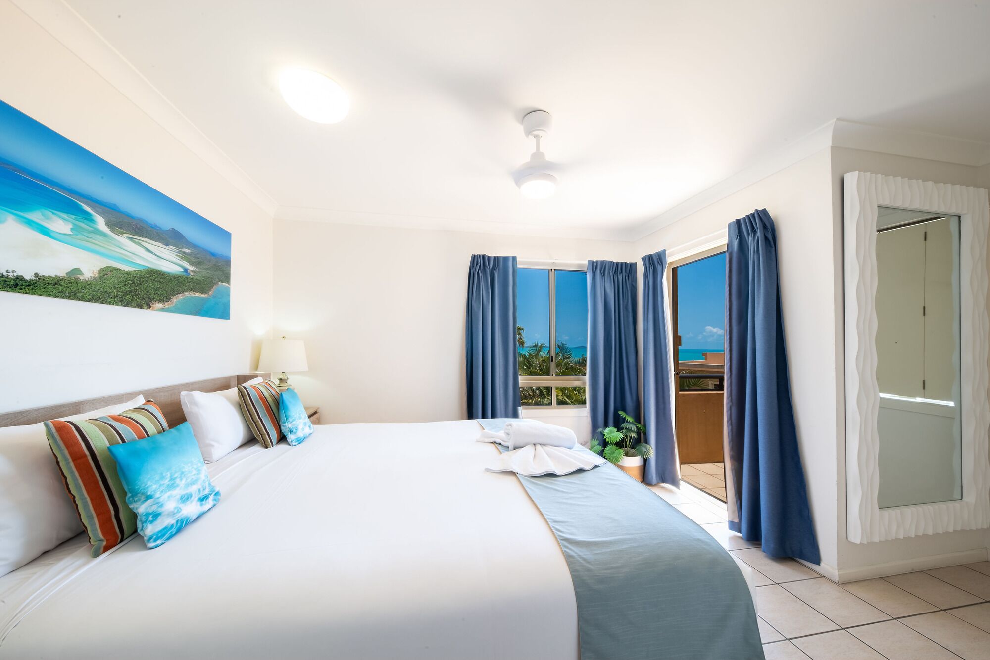 Habitación de hotel con una cama grande, un ventilador de techo, un cuadro de playa y vista al océano desde la ventana.
