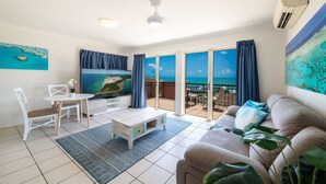 Een woonkamer met een grote televisie, een bank, een salontafel en een eethoek. Er is een balkon met uitzicht op de oceaan.