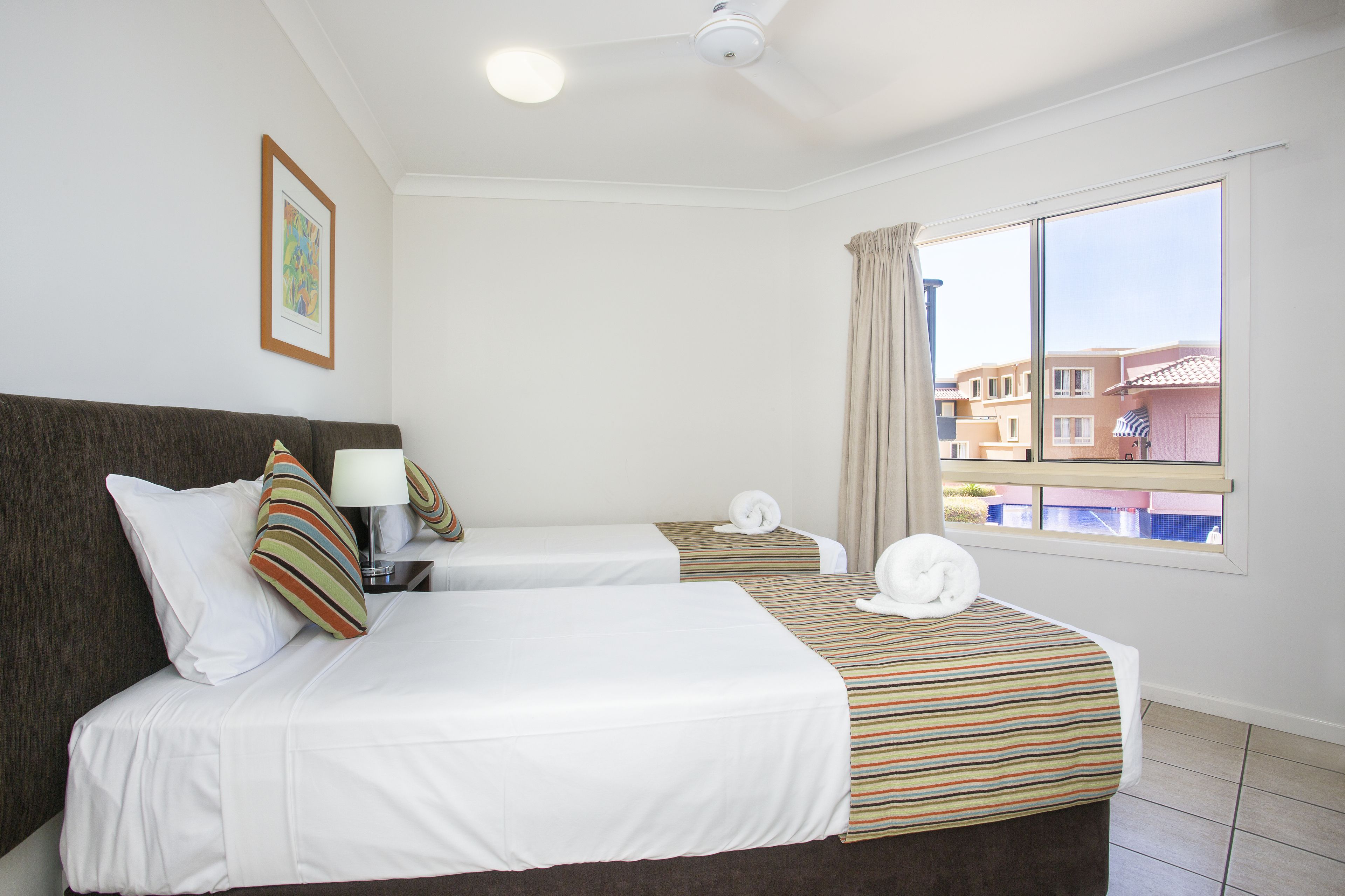 A hotel room with two beds, a ceiling fan, a window with curtains, and a view of buildings outside.
