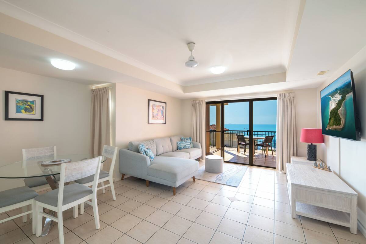 Un salon moderne avec une table à manger en verre, un canapé, un téléviseur et un balcon offrant une vue sur la mer.