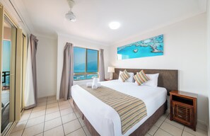 A hotel room with a large bed, a view of the ocean, a painting of a beach scene, and a wooden nightstand. - Martinique Whitsunday (Airlie Beach)