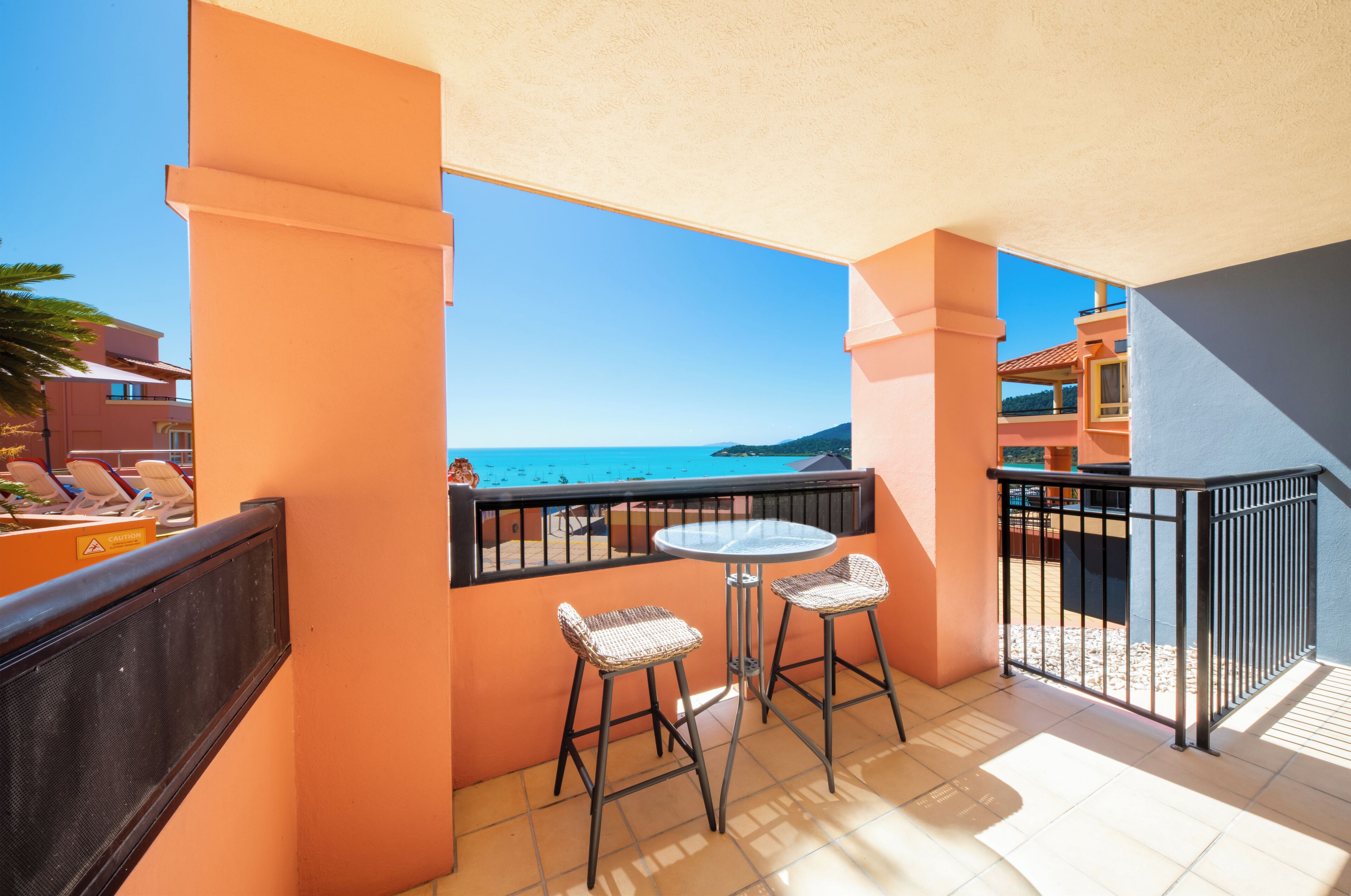 A balcony with two wicker chairs and a small table, overlooking a clear blue sea and a distant coastline.