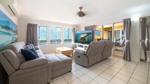 Een moderne woonkamer met een grote flatscreen televisie, beige banken, een salontafel en uitzicht op de zee door grote ramen.
