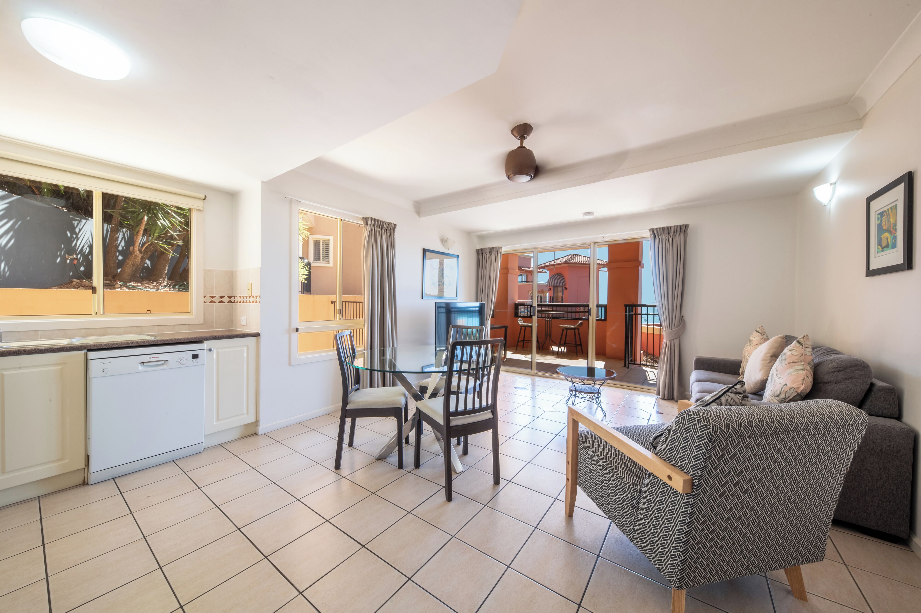 A modern kitchen with a dishwasher, dining table with chairs, a sofa, and a ceiling fan.