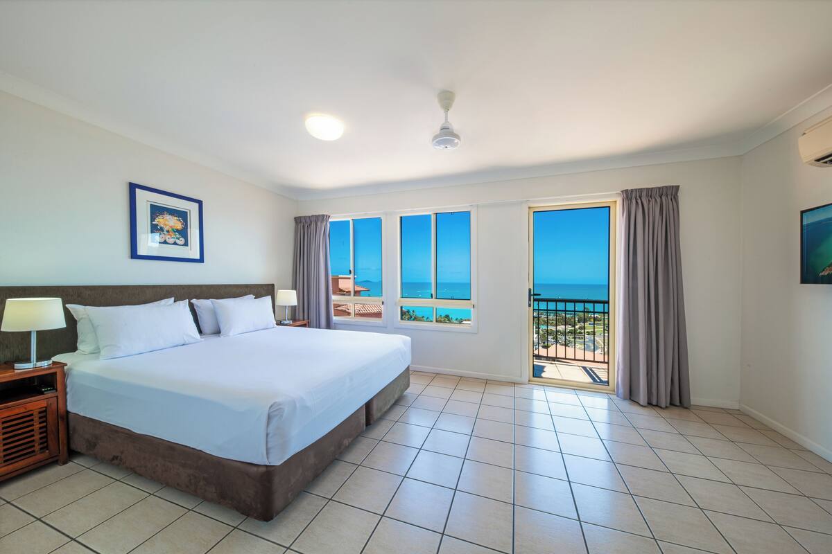 Une chambre d’hôtel avec un grand lit, une table de chevet, un ventilateur de plafond et une vue sur l’océan grâce à de grandes fenêtres.
