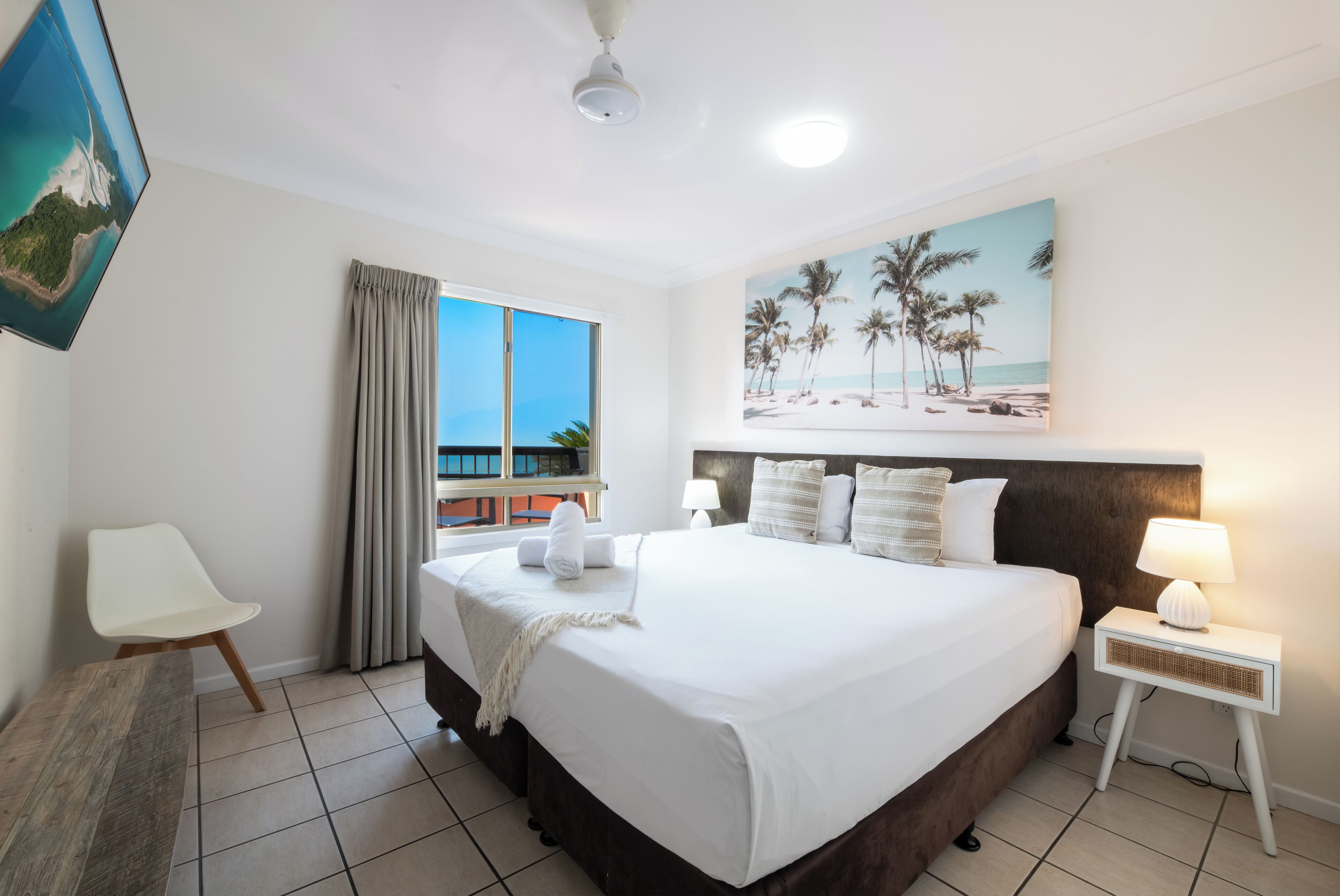 A hotel room with a large bed, a view of the sea, a painting of palm trees, and a white chair.