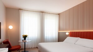 Habitación de hotel con cama, escritorio, silla, una mesita con una planta y una ventana con cortinas.