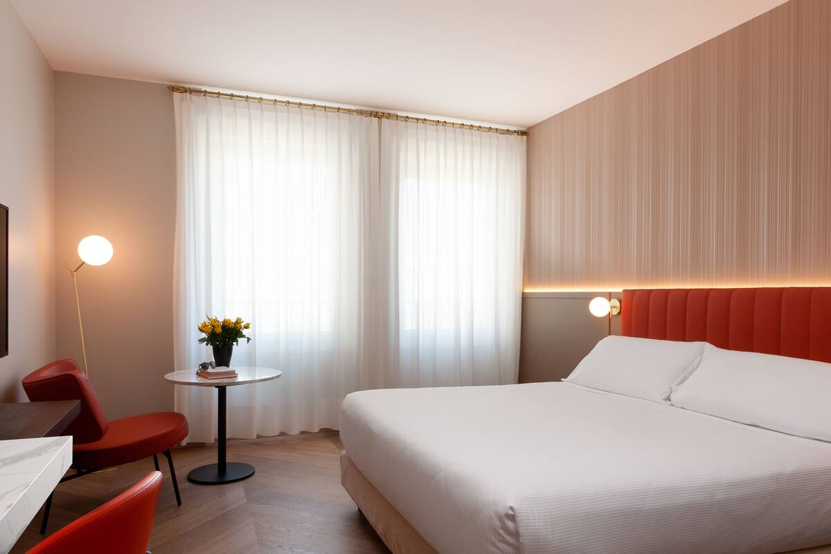 Hotelkamer met een bed, een bureau, een stoel, een kleine tafel met een plant en een raam met gordijnen.