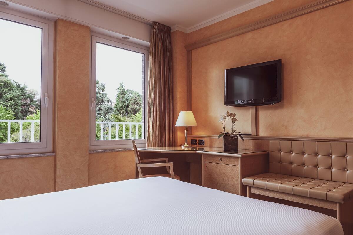 Een hotelkamer met een bed, een bureau met een lamp, een televisie en uitzicht op groen buiten.