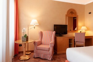 A hotel room with a bed, a desk with a television, two striped chairs, a small table with a plant, and two wall lamps.