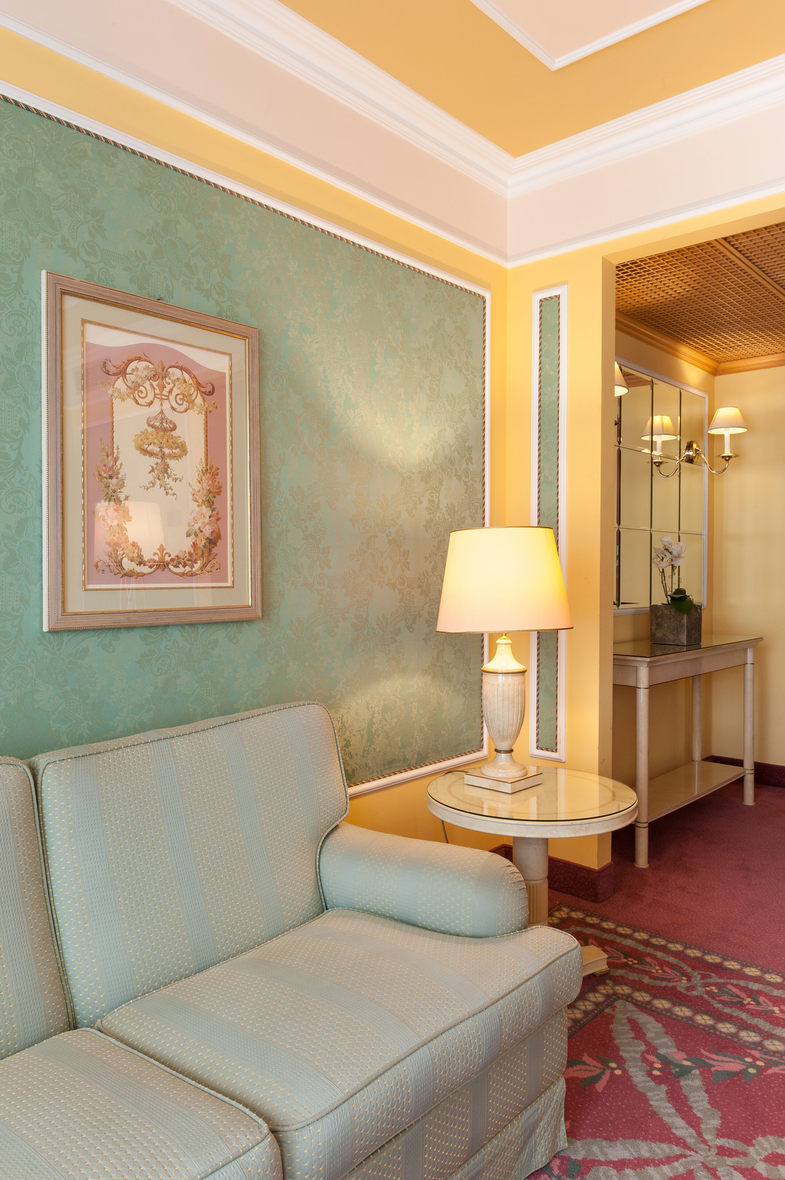 Une pièce avec un canapé bleu clair, une table ronde surmontée d’une lampe, un tableau encadré accroché au mur et un miroir au-dessus de la table.