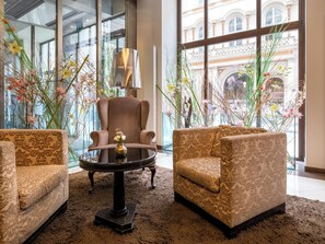 A room with a large window, a brown armchair, two patterned sofas, a round table with a vase, and a lamp. - Steigenberger Icon Herrenhof (Vienna)