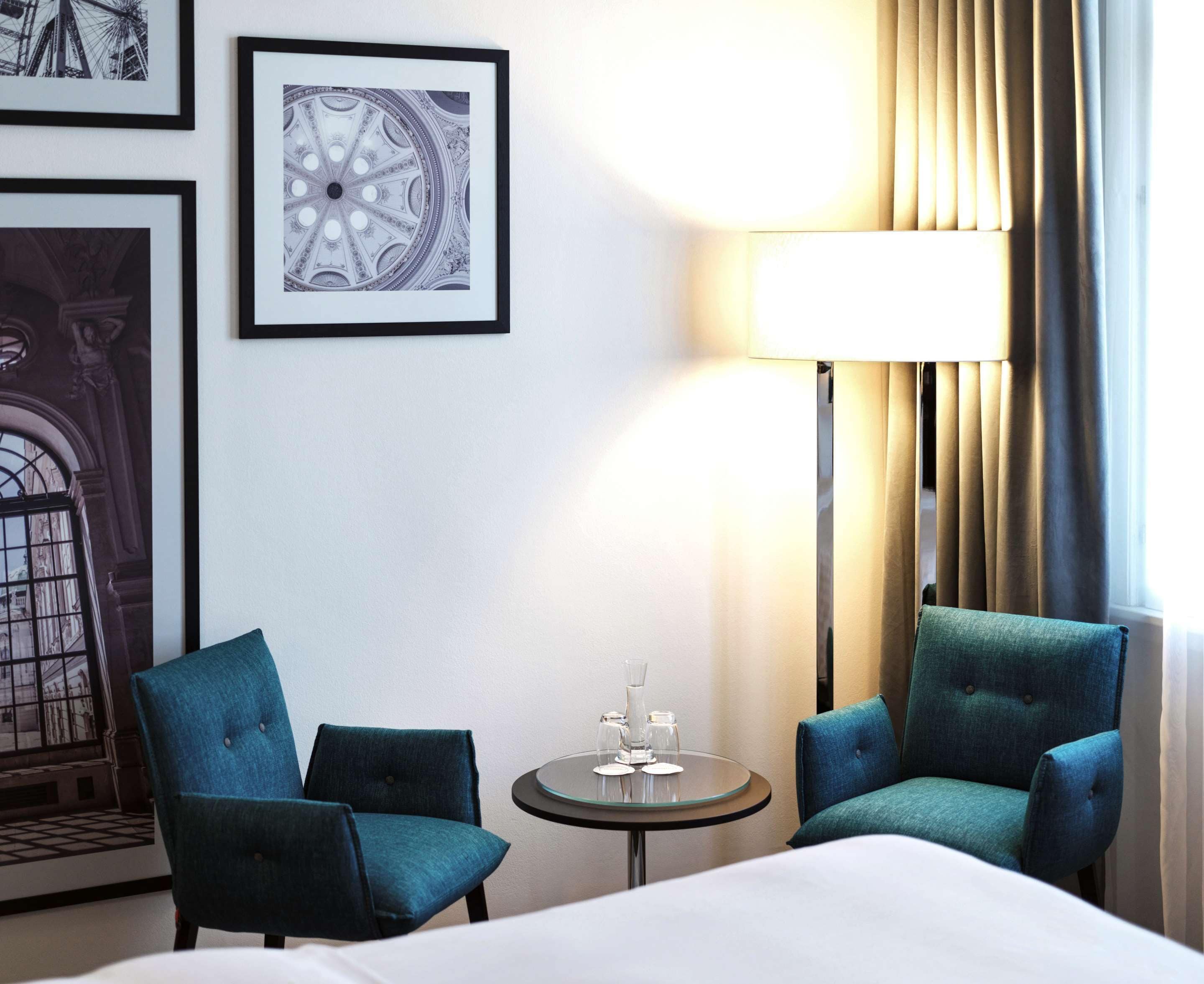A hotel room with a bed, two teal armchairs, a small round table with a glass and a vase, and framed pictures on the wall.