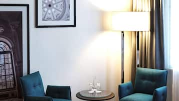 Quarto de hotel com uma cama, duas poltronas verde-água, uma mesinha redonda com um copo e um vaso, e quadros nas paredes.