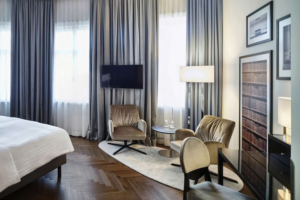 Una habitación de hotel con una cama grande, televisión, zona de estar con sillones y una estantería.