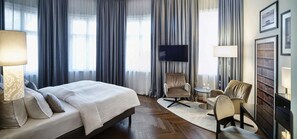 Una habitación de hotel con una cama grande, televisión, zona de estar con sillones y una estantería.
