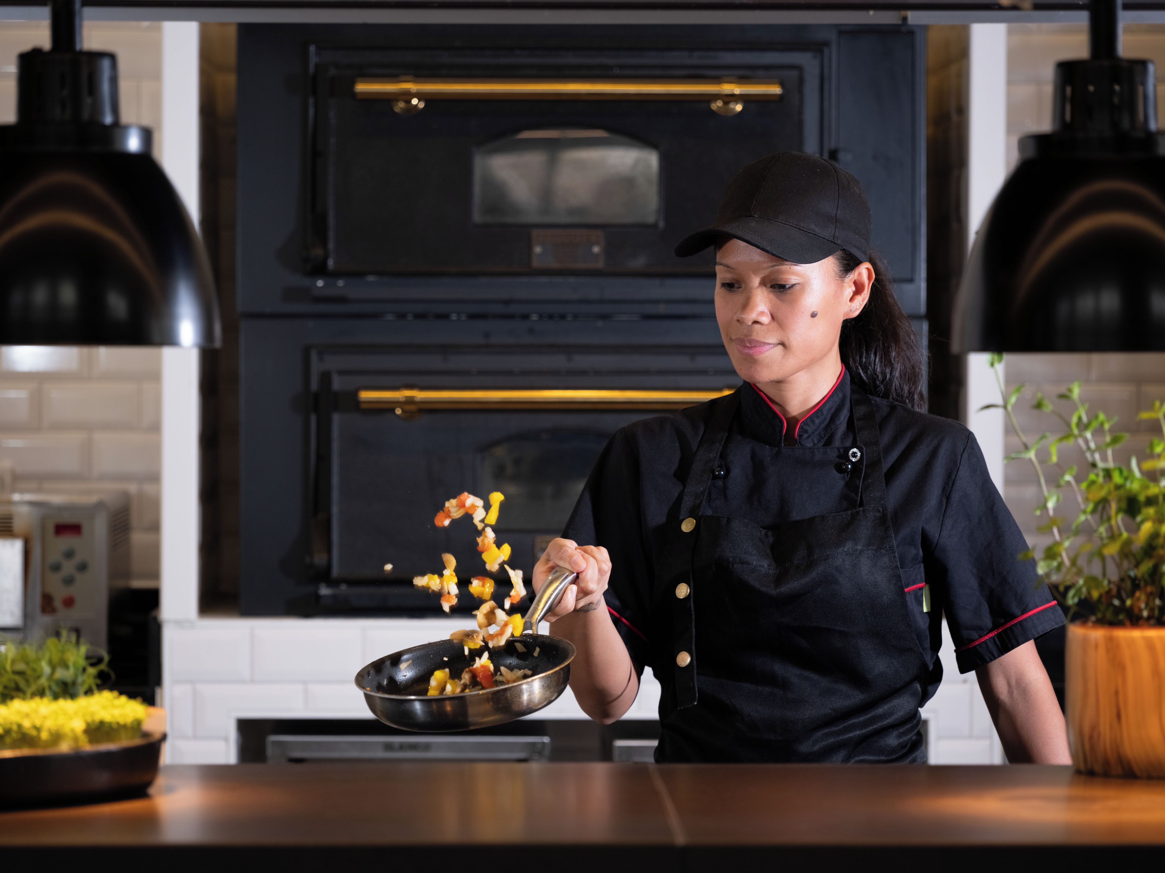 Cozinheiro de uniforme preto e touca preta, segurando uma panela com ingredientes.