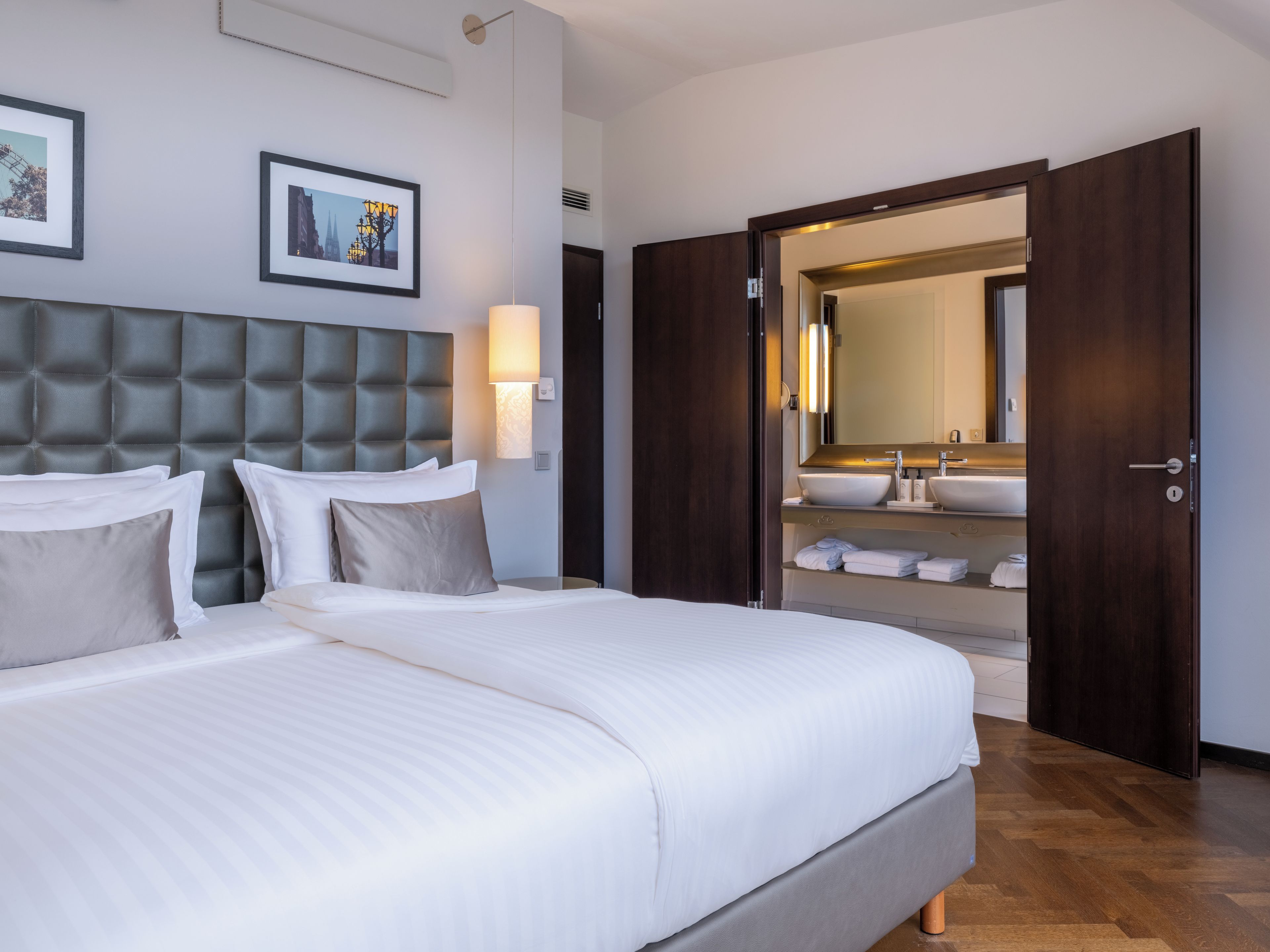 Camera d'albergo con un letto grande, una testiera, un comodino, una lampada a parete, uno specchio e un bagno con lavandino e porta asciugamani.
