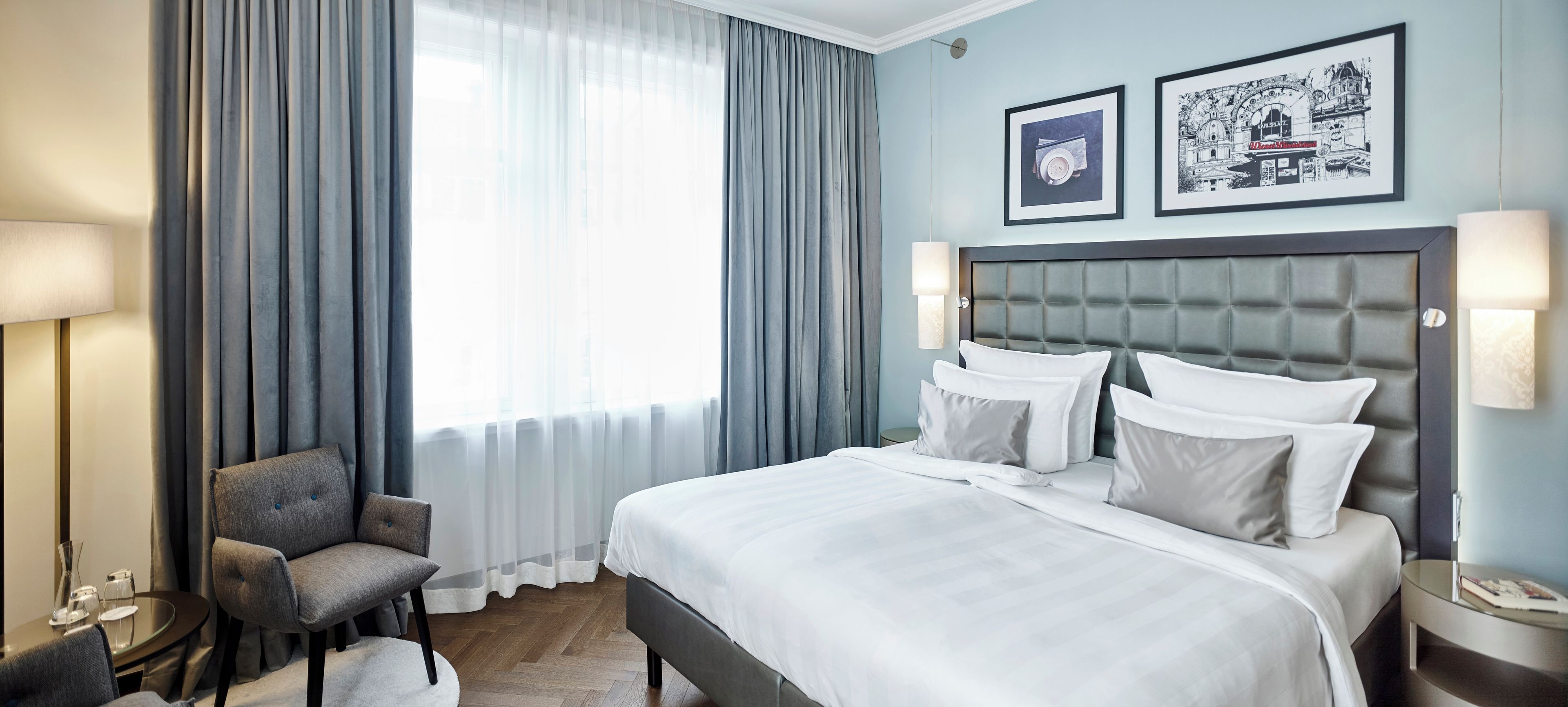Quarto de hotel com cama, poltrona, mesa de cabeceira, luminária e janela com cortinas.