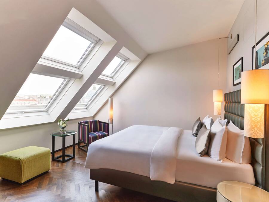 A modern hotel room with a large bed, skylight windows, a small side table, a green ottoman, and a chair.