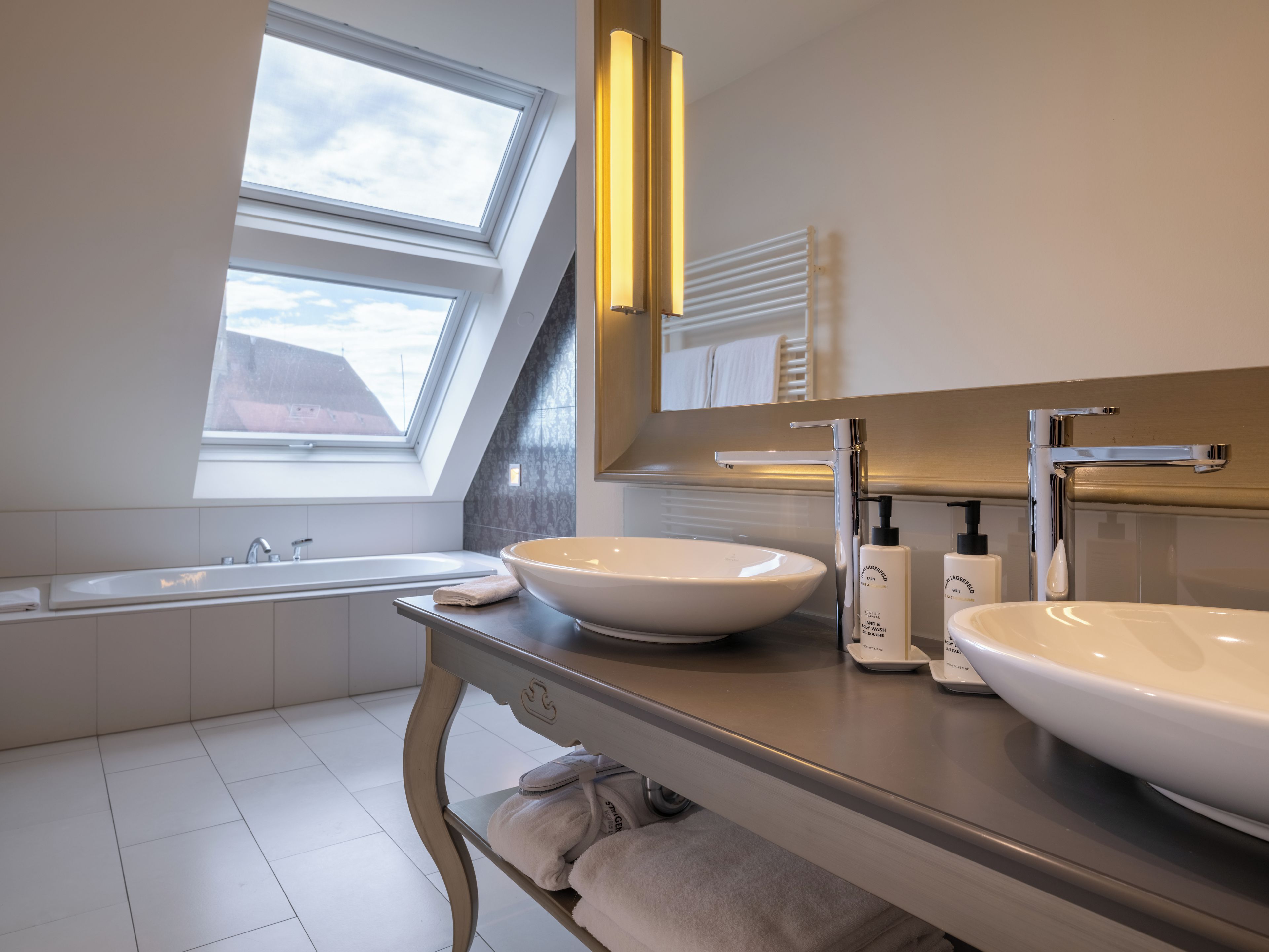 A modern bathroom with a skylight, two sinks, a bathtub, and a vanity with towels.