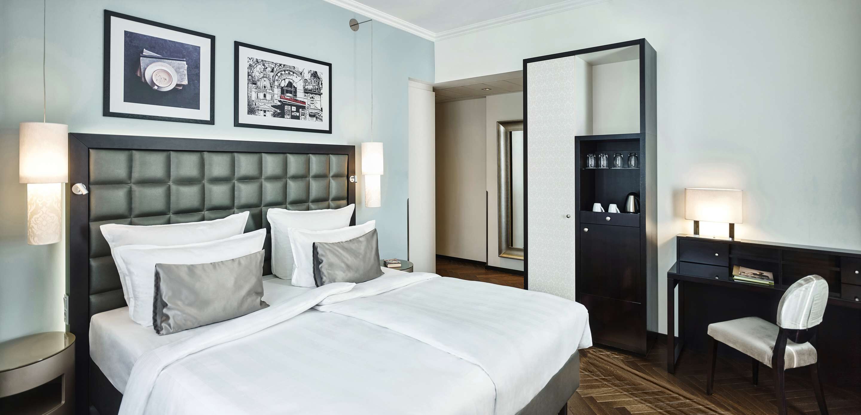 A hotel room with a large bed, two bedside tables with lamps, a desk with a chair, and a wardrobe.