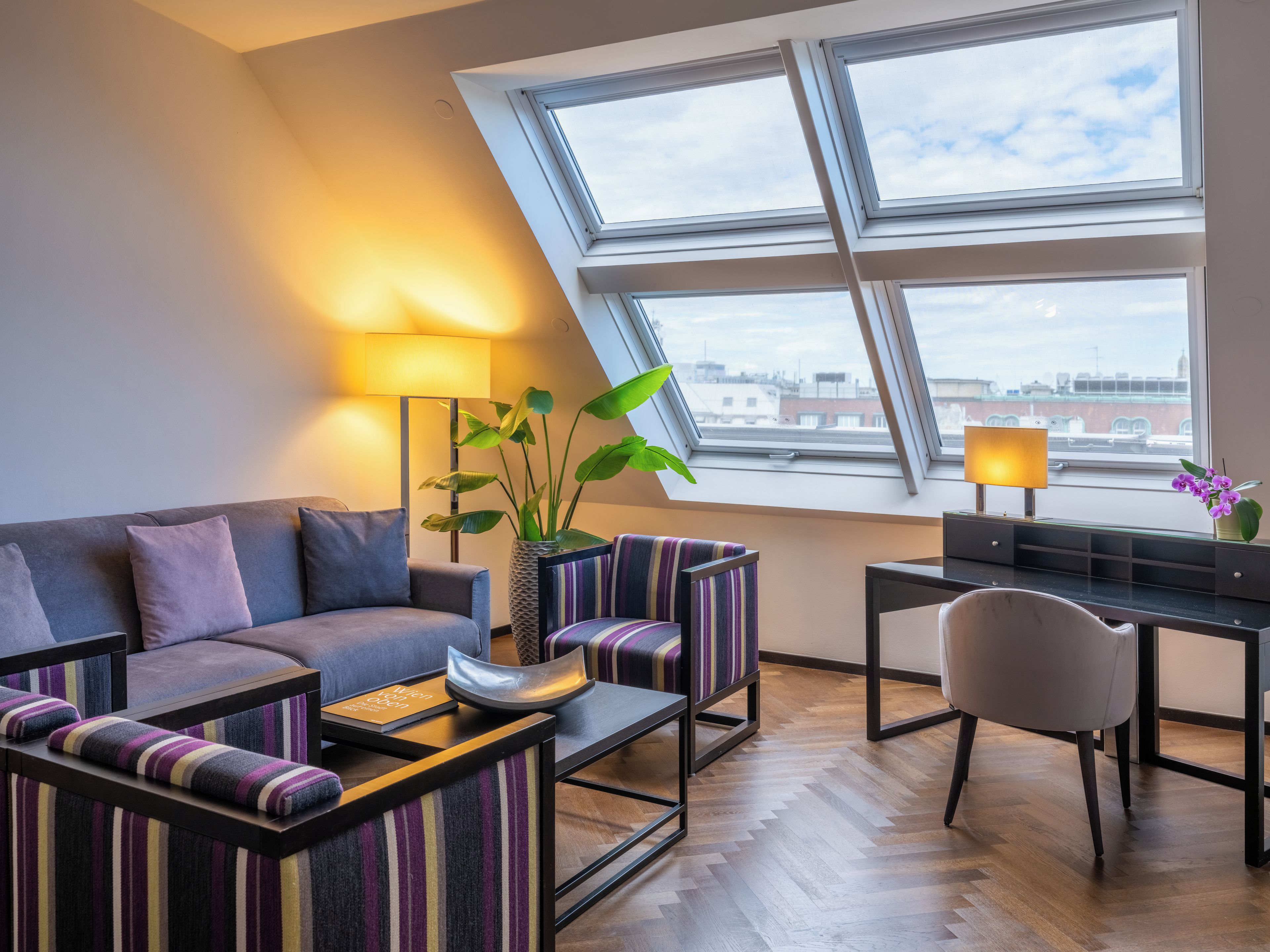 Un salon moderne comprenant un canapé, des fauteuils, une table basse et un bureau avec une lampe. Une grande fenêtre offre une vue sur le paysage urbain.