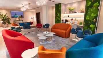 Un espace de détente moderne avec des sièges aux couleurs vives, un bar à café et un téléviseur diffusant des images d’un paysage urbain.