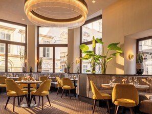 L’intérieur d’un restaurant avec des tables rondes, des chaises jaunes, de grandes fenêtres et un luminaire décoratif au plafond.