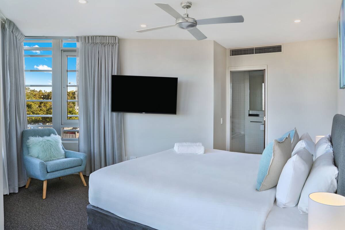 A hotel room with a large bed, a flat-screen TV mounted on the wall, a ceiling fan, and a view of the outdoors through the window.