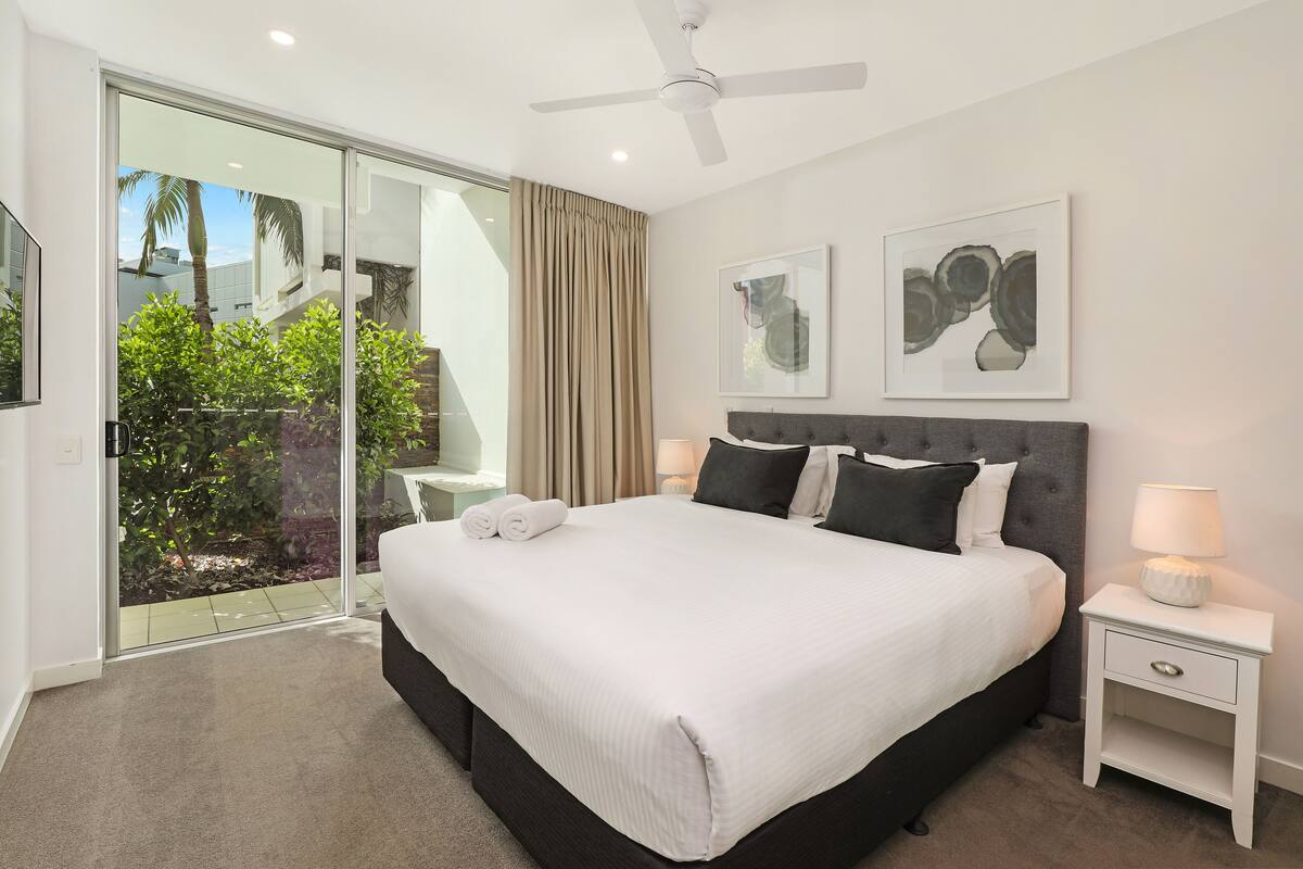 A modern hotel room with a large bed, a ceiling fan, a sliding glass door to a balcony, and two framed artworks on the wall.