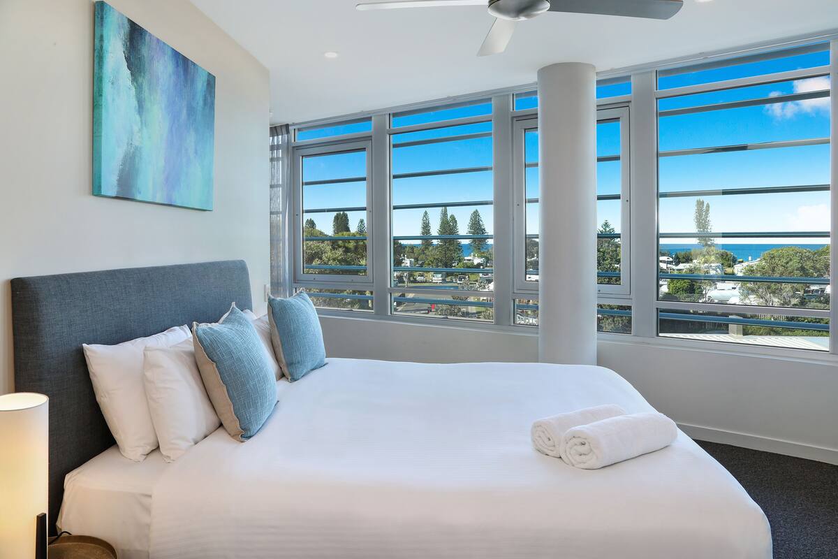 A hotel room with a large bed, a ceiling fan, and a view of the city through large windows.