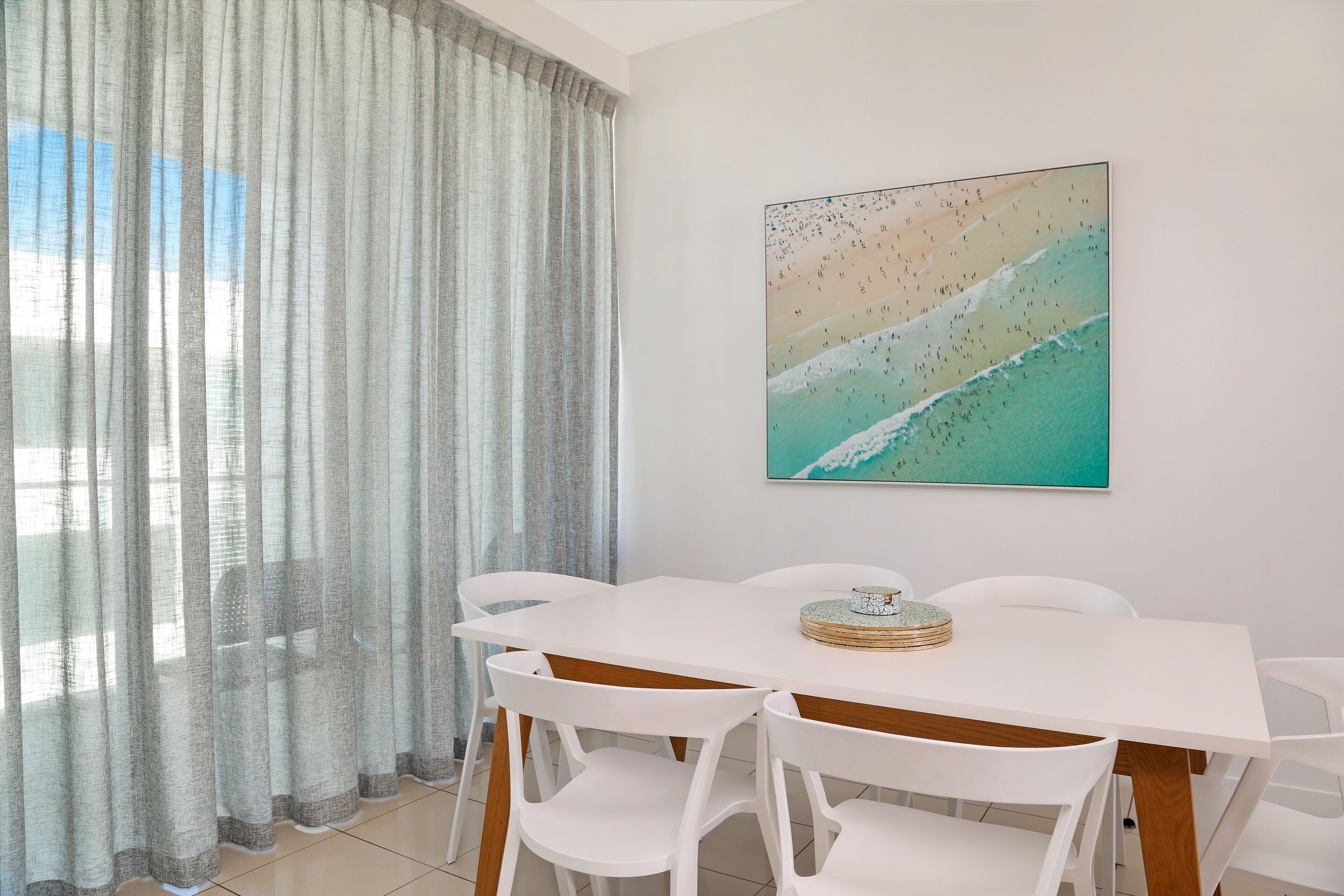 A dining area with a white table and chairs, a beach-themed painting on the wall, and sheer curtains.