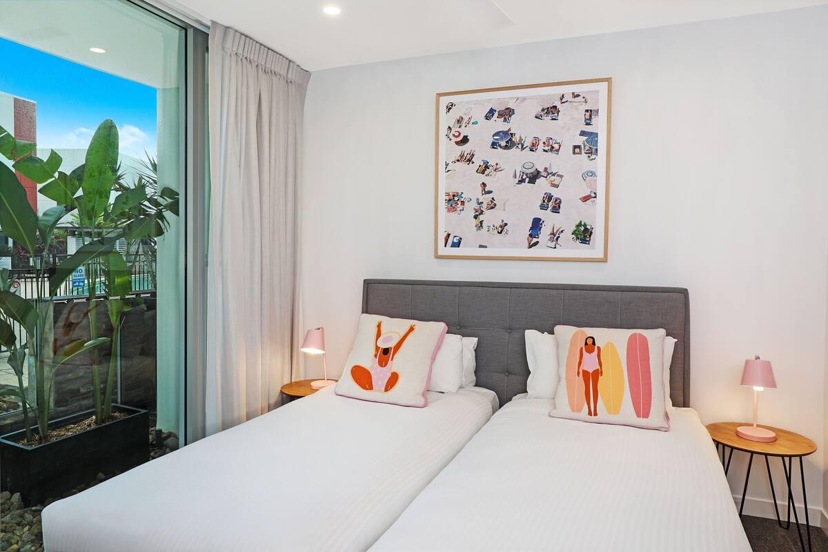 A hotel room with two beds, a large window with curtains, a wall-mounted artwork, and a small table with a lamp.